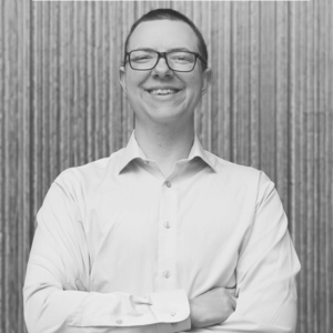 A professional photo of a white man with a white collared button up shirt smiling with his arms crossed against a gray vertical striped background