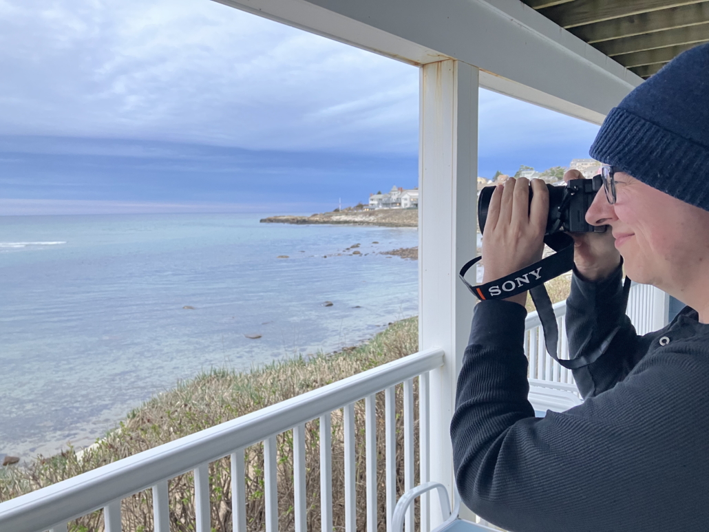 Leon Photography A young man holding a professional camera to his eye standing on a balcony taking photos of the ocean